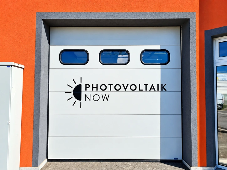 Weißes Garagentor mit schwarzer Photovoltaik-Firmenbeschriftung und Logo auf orangefarbener Fassade.