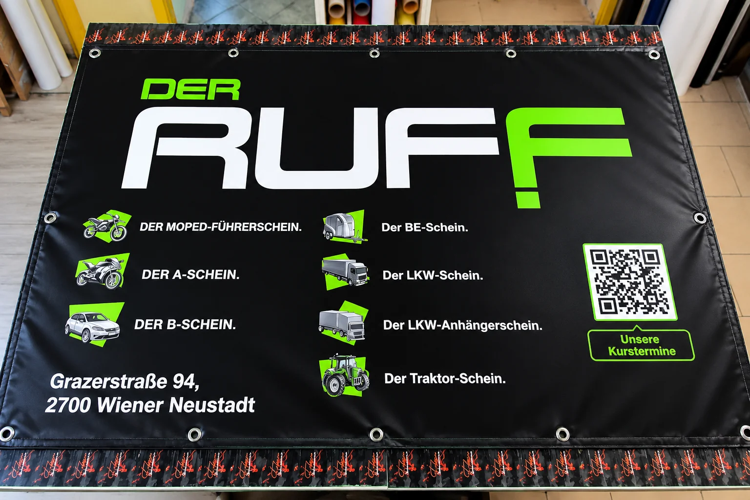 Bedrucktes schwarzes Werbebanner mit Führerscheinklassen, Fahrzeug-Symbolen, QR-Code und Adresse in Wiener Neustadt.